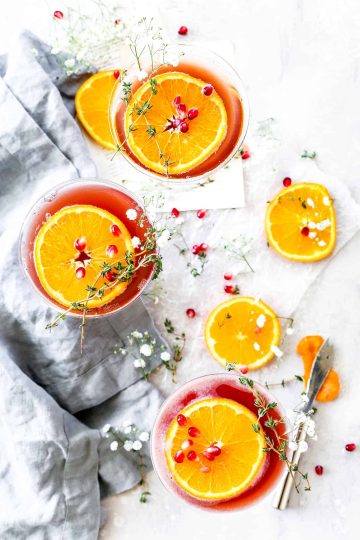 Maroon mocktails in martini glasses garnished with fresh thyme sprigs, pomegranate seeds and orange slices.