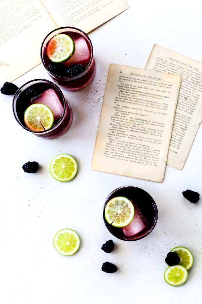 Overhead shot of three Vigilante Shit mocktails garnished with lime and blackberries on a white surface with scattered book pages.