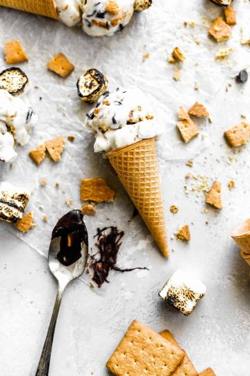 S'mores ice cream cone lying flat on a white surface next to a spoon covered in chocolate and graham cracker chunks.