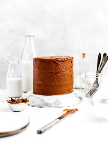 Vegan chocolate cake, a jar of forks, jug of milk and a knife covered in chocolate frosting.