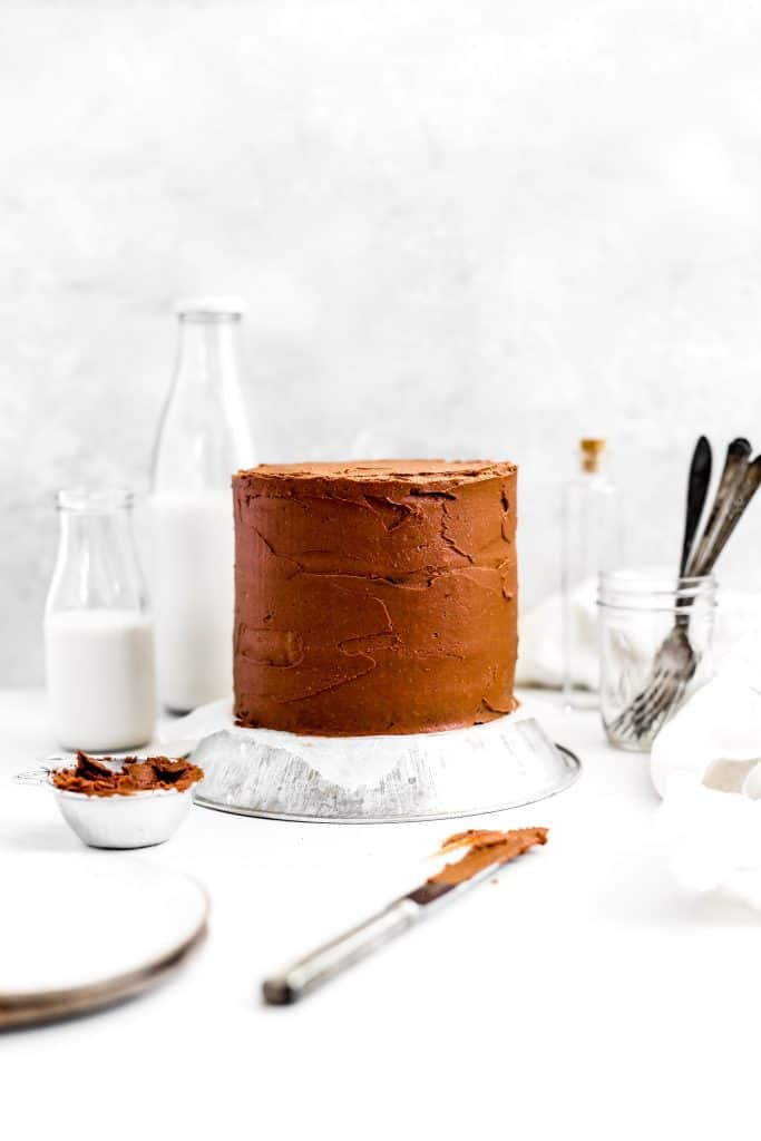 Vegan chocolate cake, a jar of forks, jug of milk and a knife covered in chocolate frosting.