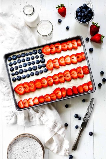 Vegan flag cake in a silver pan, a cup of milk, a cup of blueberries, a stack of white plates and a knife covered in frosting.