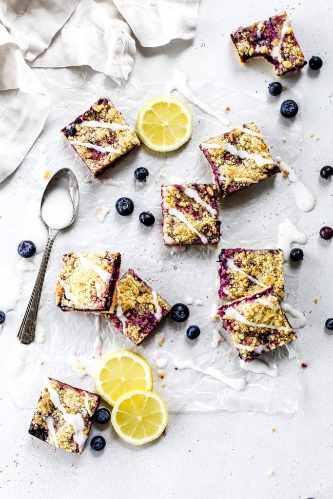 Vegan lemon blueberry bars scattered on a white surface drizzled in lemon glaze surrounded by fresh blueberries.