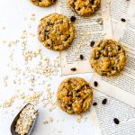 Vegan oatmeal raisin cookies on a white surface with loose book pages and a scoop of rolled oats.