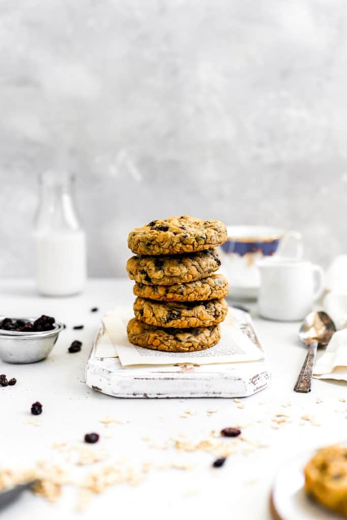 Stack of vegan oatmeal raisin cookies on a white wooden cutting board in front of a jug of milk and a cup of tea.