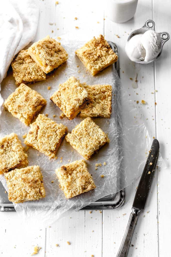 Vegan peach crumble bars on a baking sheet next to a cup of vanilla ice cream, a jug of milk and a knife.