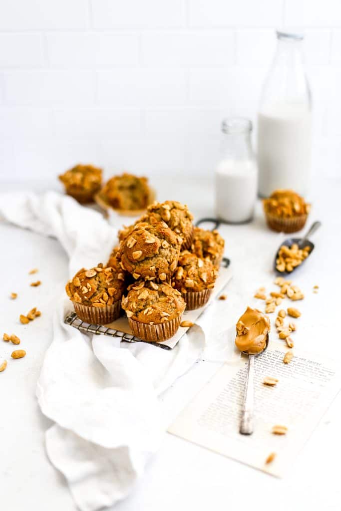 Vegan peanut butter banana muffins on a wire cooling rack, a scoop of peanuts a spoon full of peanut butter and a jug of milk.