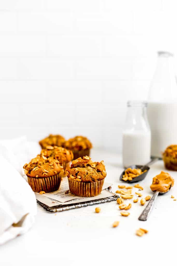 Vegan peanut butter banana muffins on a wire cooling rack, a spoonful of peanut butter, and a scoop of peanuts in front of a jug of milk.