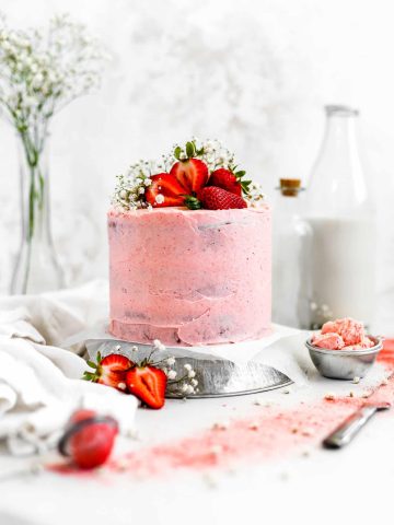 Vegan strawberry banana cake topped with strawberries in front of a jug of milk and a cup of extra strawberry frosting.