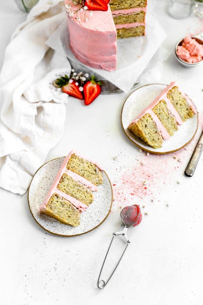 Vegan strawberry banana cake with two slices on white plates, a jug of milk and a knife covered in strawberry frosting.