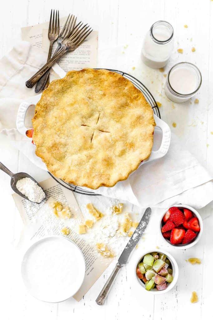 Strawberry rhubarb pie on a wire cooling rack, a stack of white plates, forks, a glass of milk, a cup of strawberries and a cup of rhubarb.