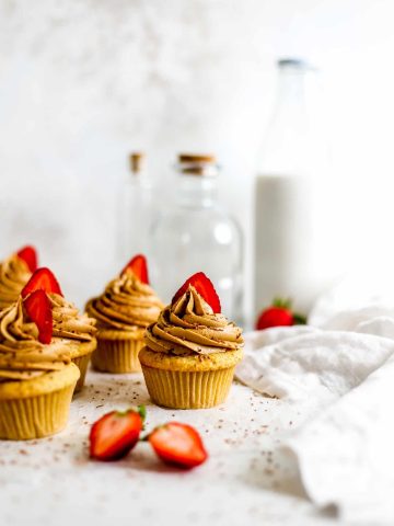 Vegan strawberry tiramisu cupcakes on a white surface in front of glass bottles and a jug of milk, next to a white linen napkin.