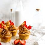 Vegan strawberry tiramisu cupcakes on a white surface in front of glass bottles and a jug of milk, next to a white linen napkin.