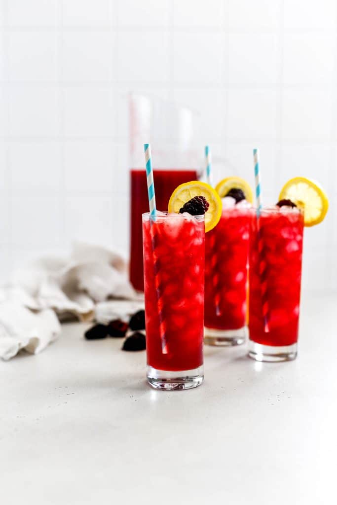 Three glasses of blackberry lemonade garnished with lemon slices and blackberries in front of a pitcher full of lemonade.