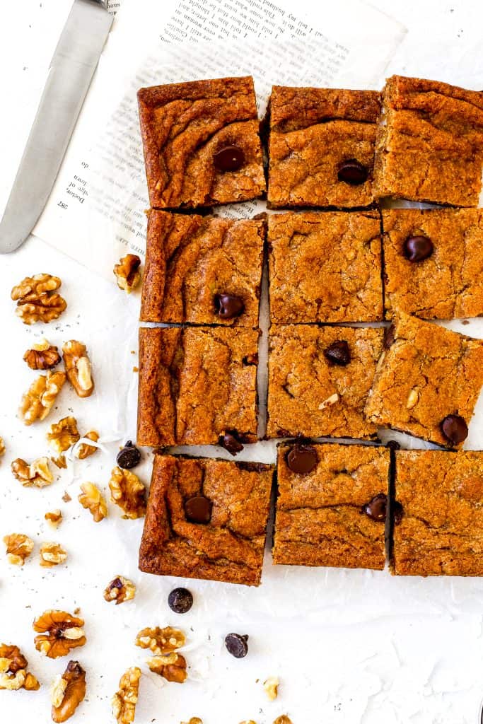 Sliced vegan blondies on a white surface, a knife and walnuts.