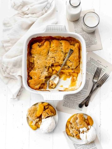 White casserole dish of vegan peach cobbler and two white plates of cobbler topped with vanilla ice cream.