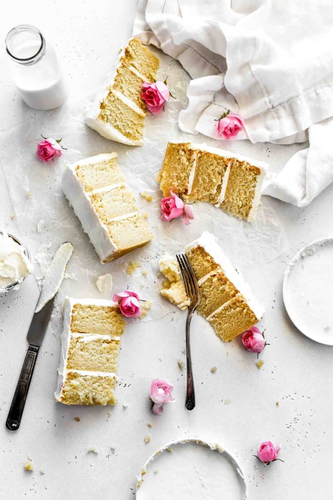 Slices of vegan vanilla cake lying directly on a white surface, a knife covered in frosting and pink flowers scattered throughout.