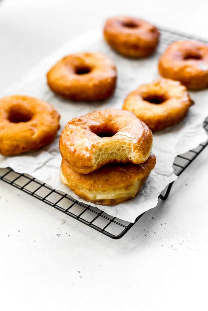 Glazed vegan donuts stacked on a wire cooling rack, one donut with a bite taken out of it.