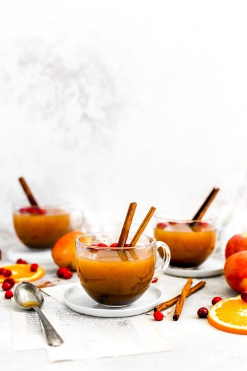 Three mugs of apple cider surrounded by orange slices, apples and cinnamon sticks.