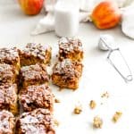 Square slices of apple cinnamon coffee cake, a jug of milk, two apples and a powdered sugar duster.