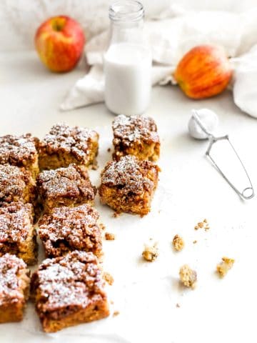 Square slices of apple cinnamon coffee cake, a jug of milk, two apples and a powdered sugar duster.