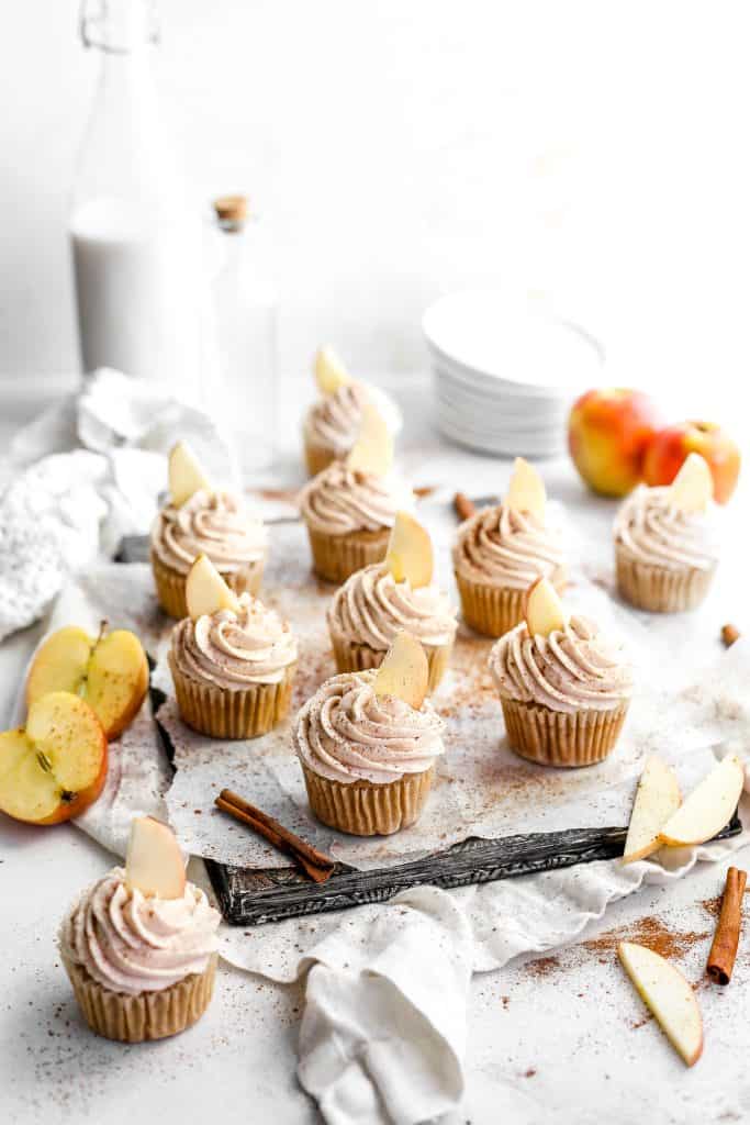 Vegan apple cinnamon cupcakes garnished with fresh apple slices sitting on a baking sheet in front of a jug of milk and a stack of white plates.