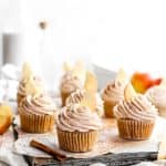 Frosted vegan apple cinnamon cupcakes on a baking sheet surrounded by apple slices and cinnamon sticks.