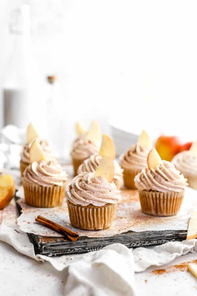 Frosted vegan apple cinnamon cupcakes on a baking sheet surrounded by apple slices and cinnamon sticks.