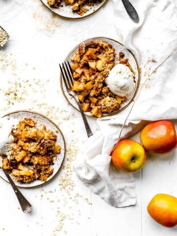 Two plates of vegan apple crisp topped with vanilla ice cream with serving forks.