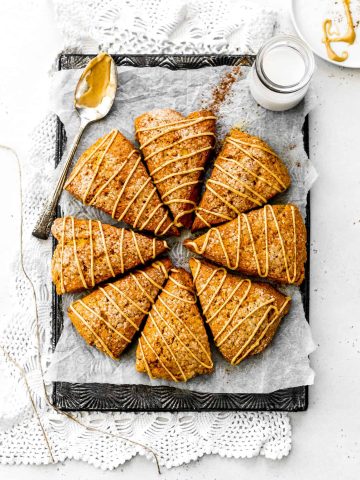 Vegan Pumpkin Scones arranged in a circle on a baking sheet drizzled in maple icing next to a glass of milk and a spoon covered in glaze.