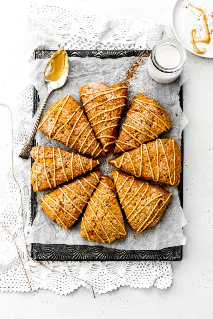 Vegan Pumpkin Scones arranged in a circle on a baking sheet drizzled in maple icing next to a glass of milk and a spoon covered in glaze.