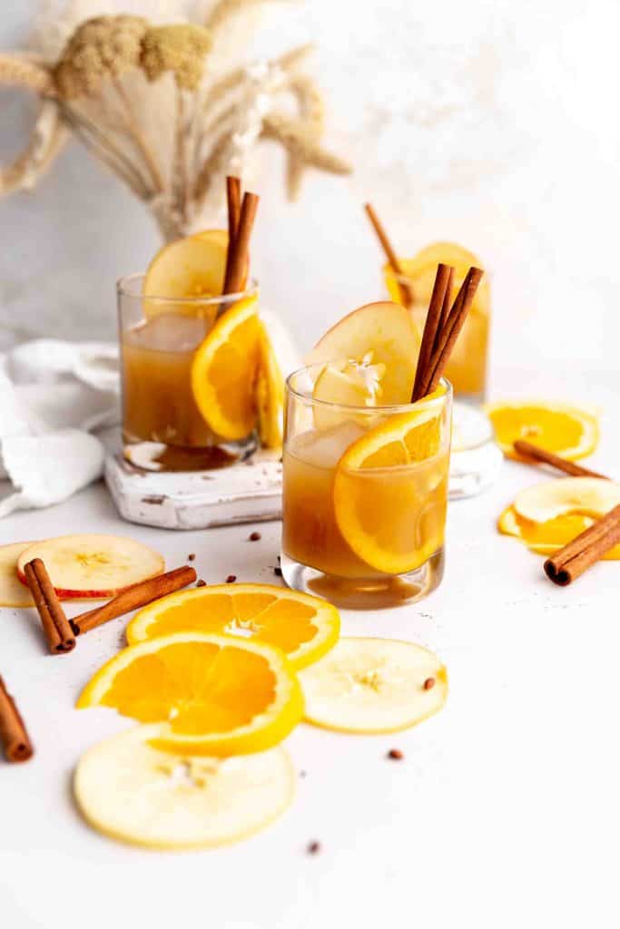 Three apple cider daiquiris on a wooden cutting board surrounded by orange slices, apple slices and cinnamon sticks.