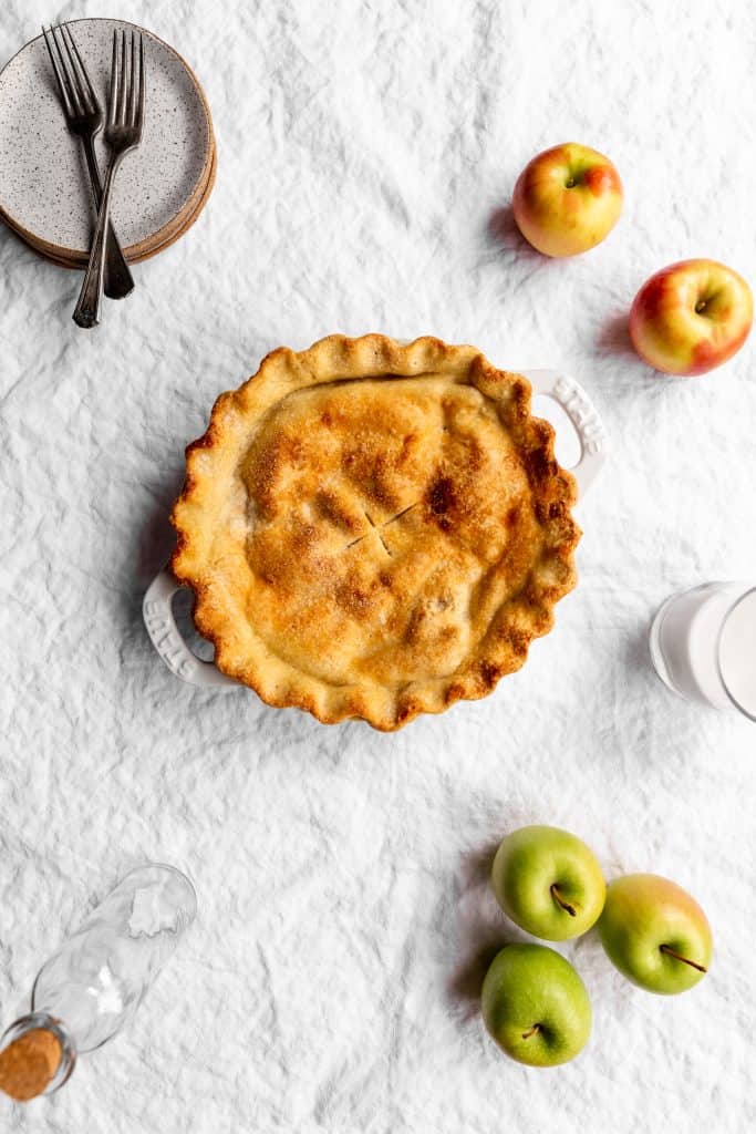 Vegan apple pie surrounded by apples, a glass of milk and a stack of white plates.