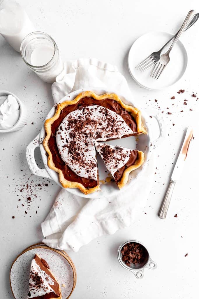 Vegan chocolate fudge pie with a slice missing surrounded by a stack of white plates, forks, a glass of milk, a cup of whipped cream and a slice of the pie on a plate.