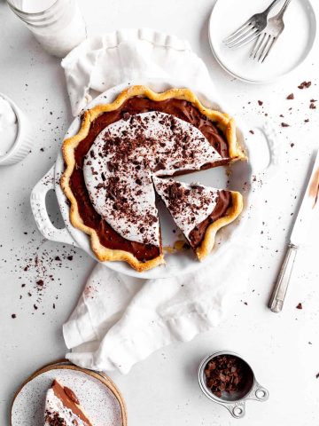 Vegan chocolate fudge pie with a slice missing surrounded by a stack of white plates, forks, a glass of milk, a cup of whipped cream and a slice of the pie on a plate.