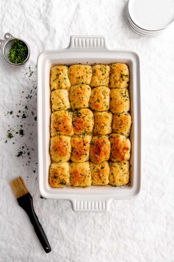 White baking dish holding baked vegan butter rolls next to a pastry brush, a stack of white plates and a cup of fresh parsely.