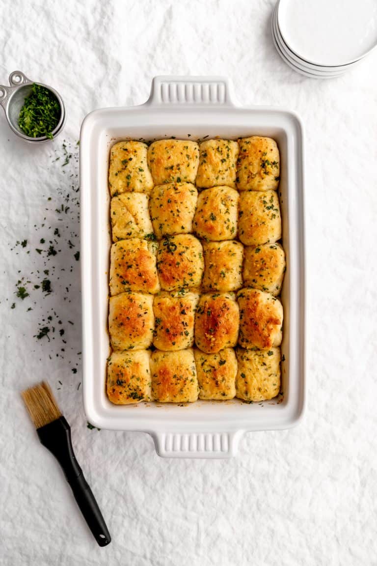 White baking dish holding baked vegan butter rolls next to a pastry brush, a stack of white plates and a cup of fresh parsely.