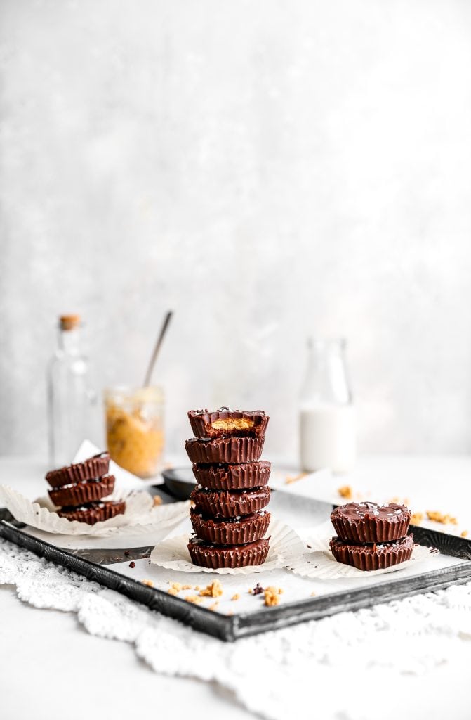 Vegan peanut butter cup with a bite taken out of it stacked on top of more peanut butter cups in front of a jar of peanut butter and a jug of milk.