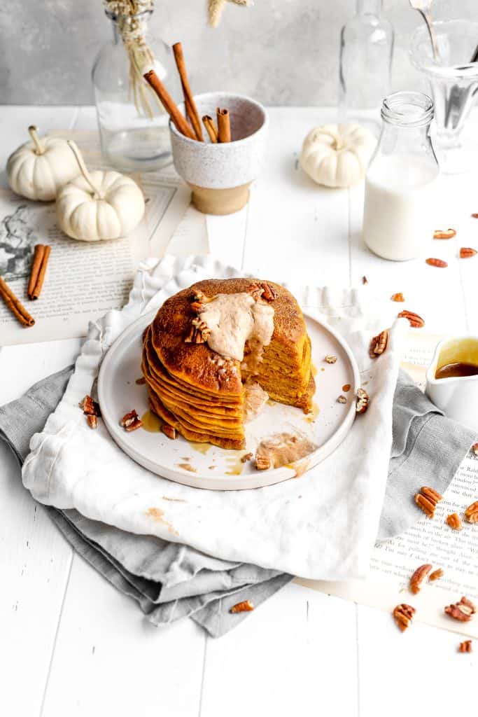 Stack of vegan pumpkin pancakes with a triangle cut out topped with oozing, cinnamon butter in front of a cup of cinnamon sticks, white pumpkins and a jug of milk.