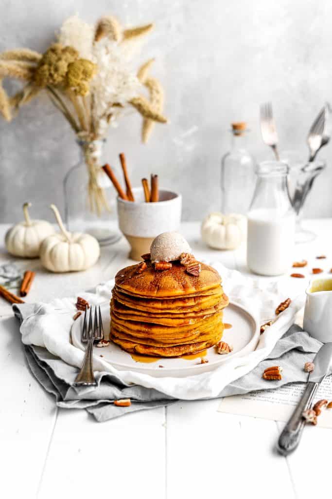 Stack of vegan pumpkin pancakes drizzled in maple syrup and topped with a scoop of cinnamon butter in front of a jug of milk and a cup of cinnamon sticks.