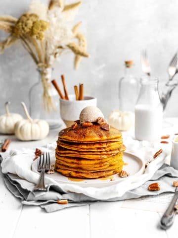 Stack of vegan pumpkin pancakes drizzled in maple syrup and topped with a scoop of cinnamon butter in front of a jug of milk and a cup of cinnamon sticks.