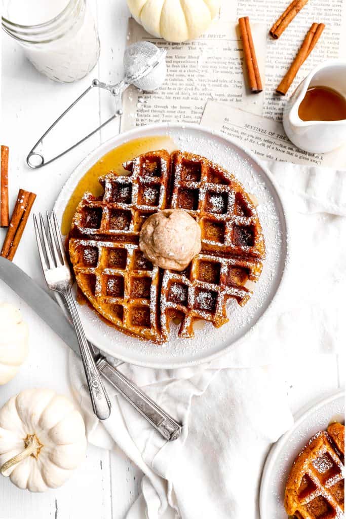 Close up of a vegan pumpkin waffle on a white plate topped with maple syrup, cinnamon butter and powdered sugar.