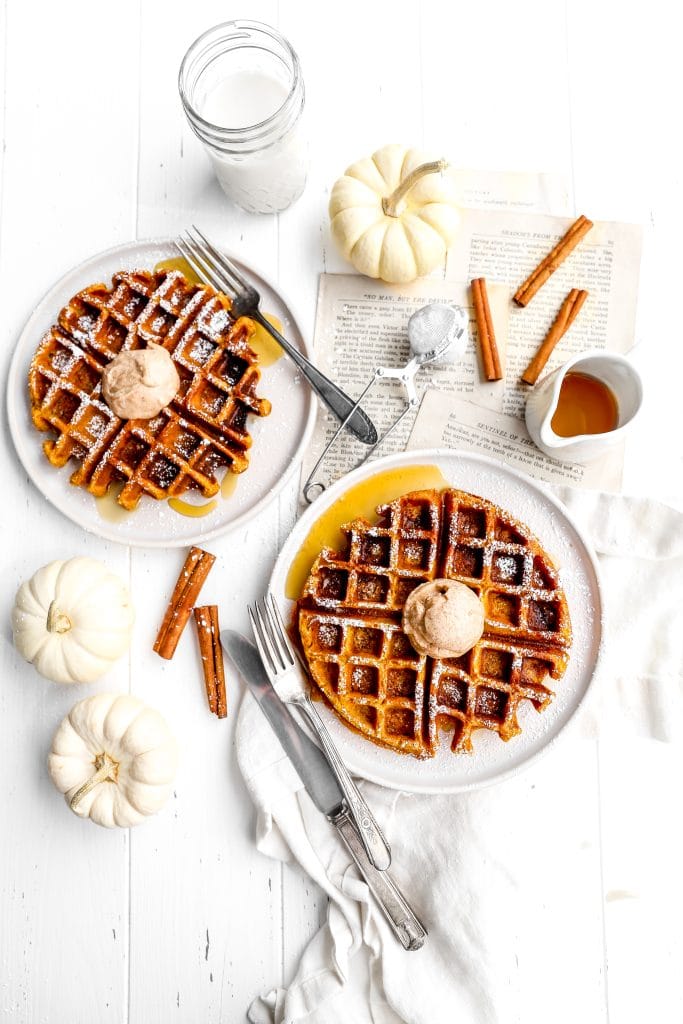 Two vegan pumpkin waffles on plates next to a glass of milk, cinnamon sticks and white pumpkins.