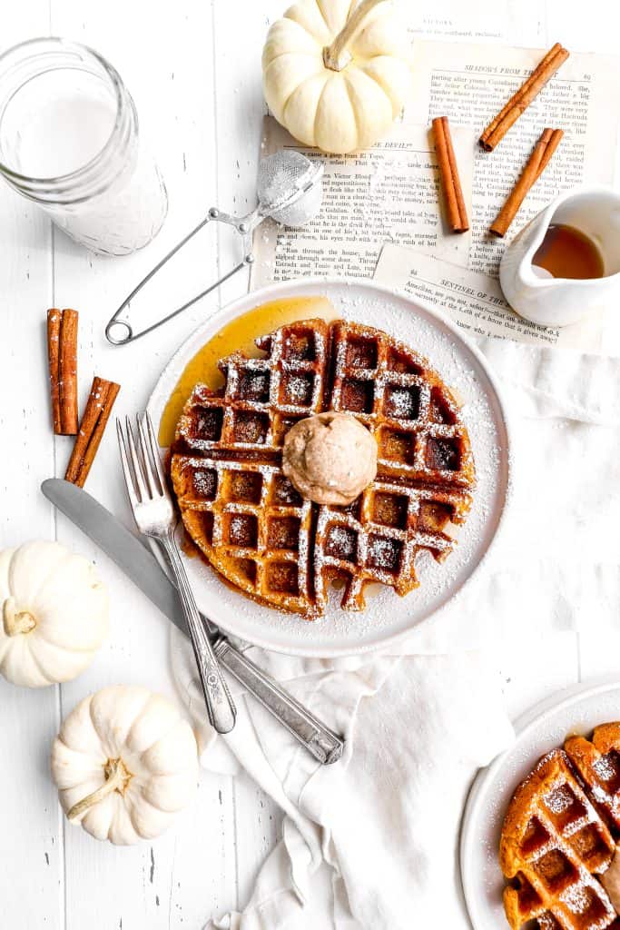 Vegan pumpkin waffle on a white plate with a knife and fork circled by a glass of milk, a linen napkin, cinnamon sticks and a cup of maple syrup.