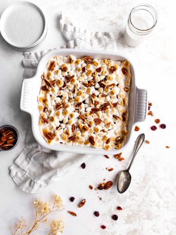 Vegan sweet potato casserole sitting on top of a white linen surrounded by a glass of milk, a stack of white plates, a rustic spoon and a cup of pecans.