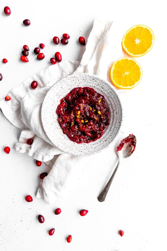 Orange cranberry sauce in a white bowl sitting on a linen napkin surrounded by fresh cranberries and orange slices.