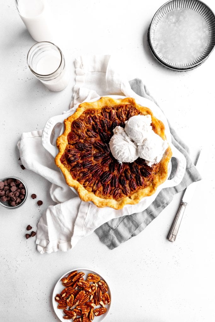 Vegan chocolate bourbon pecan pie topped with scoops of vanilla ice cream next to a jug of milk and a stack of silver plates.