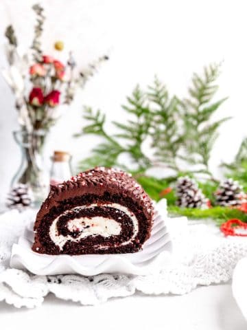 Vegan chocolate peppermint yule log on a white serving platter in front of a vase of roses, a jar of crushed candy canes and a few pinecones.