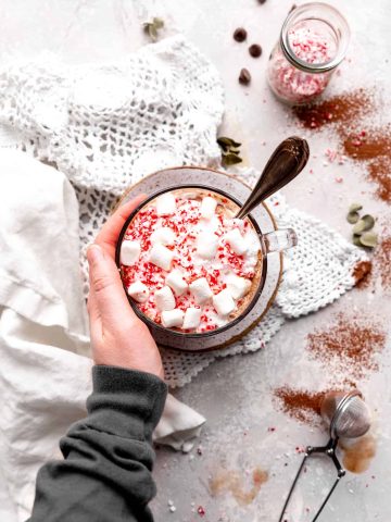 Hand wrapped around a mug of vegan peppermint hot chocolate with a spoon in it.