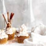 Vegan pumpkin pie cupcakes topped with whipped cream next to a white linen napkin, white pumpkins, a jar of cinnamon sticks and a jug of milk.